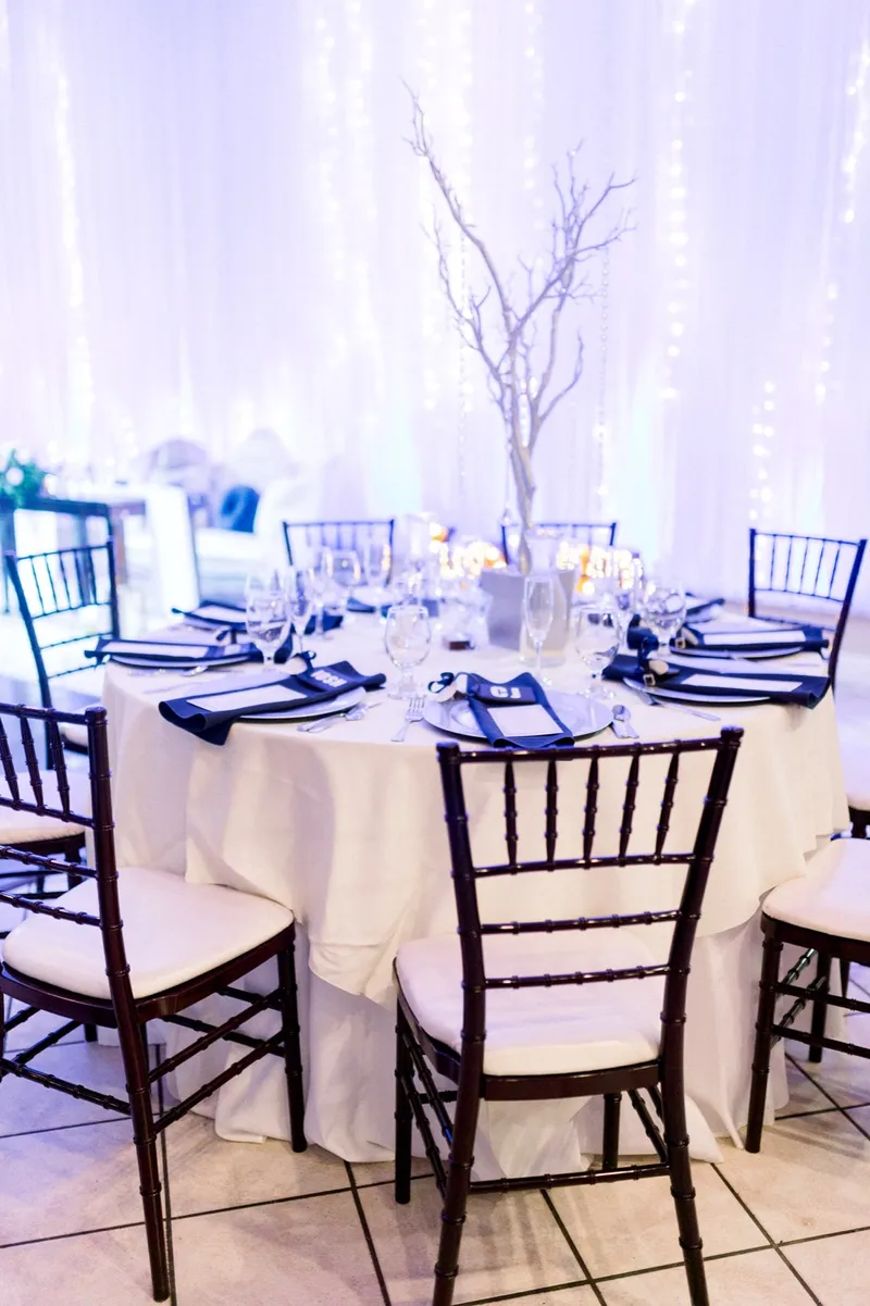 A reception table with navy napkins and a lit white branch centerpiece under the room's string lights