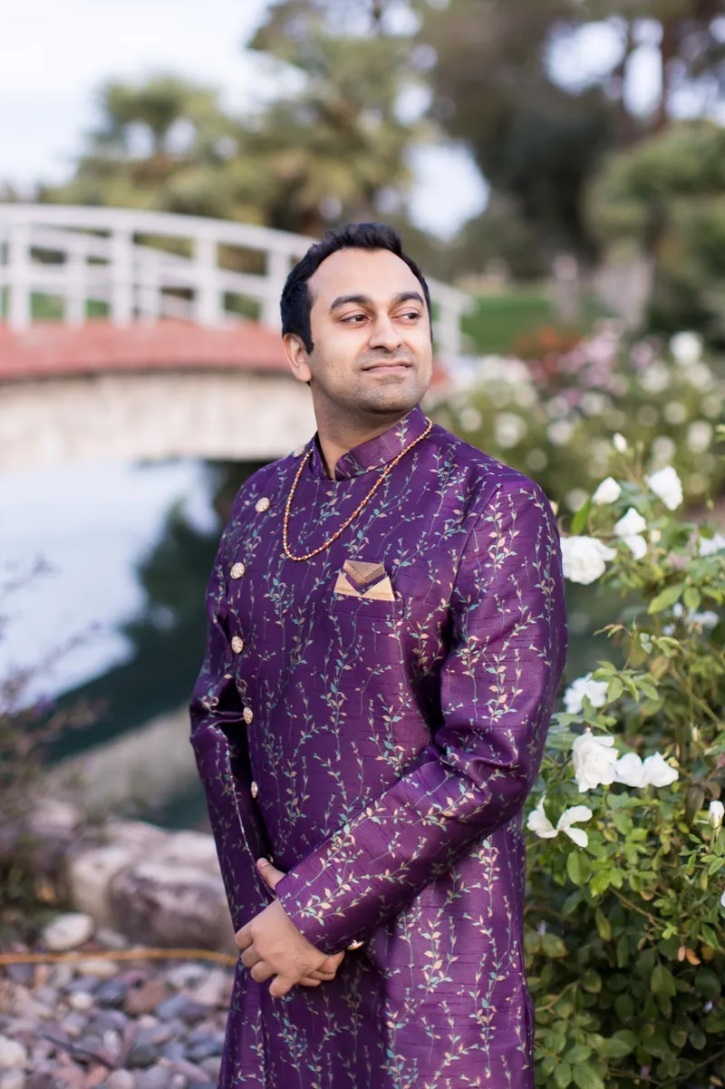 Groom in a purple embroidered sherwani standing beside the water, the white bridge and a rose bush behind him