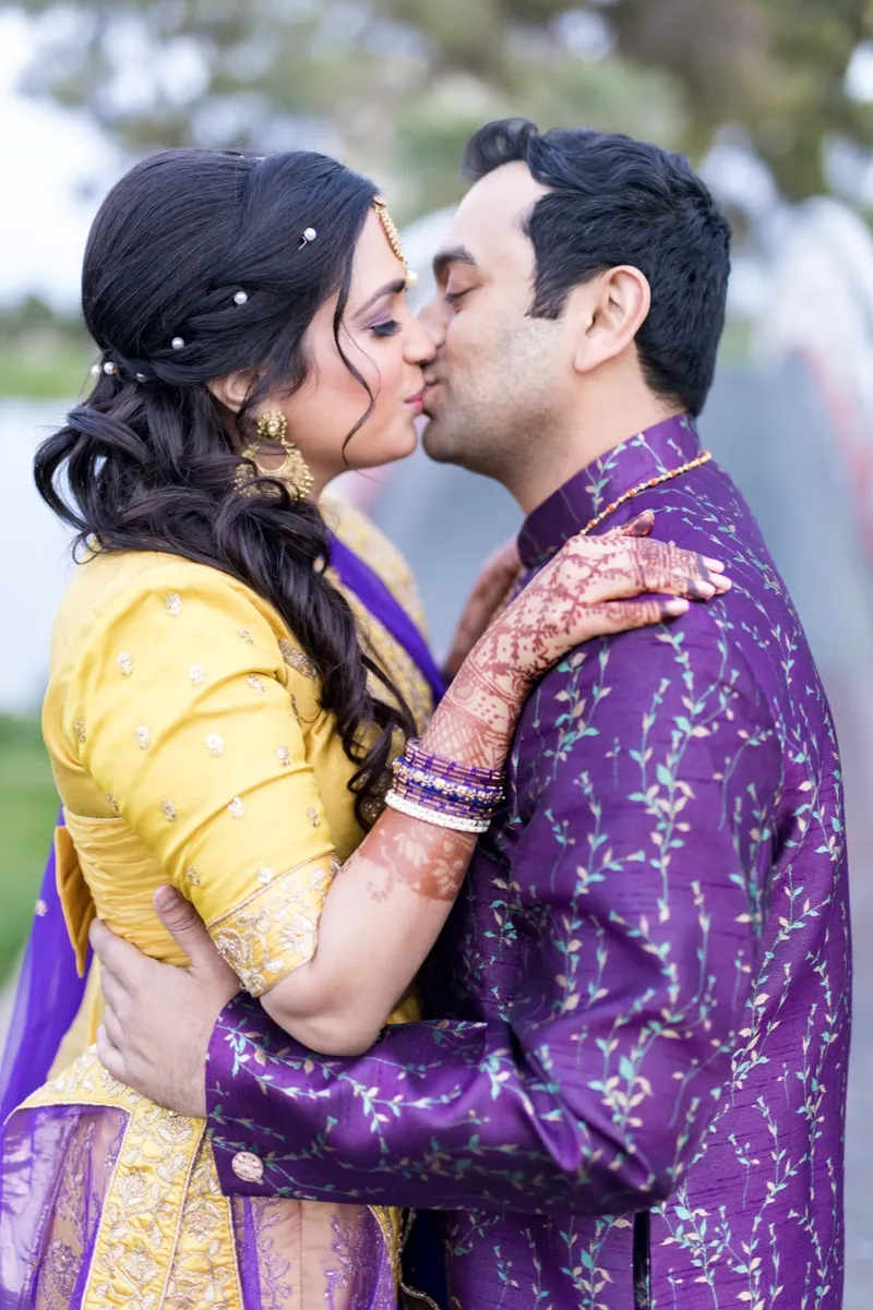 Couple kissing on the lawn, the bride in yellow and purple with henna on her hands and the groom in purple