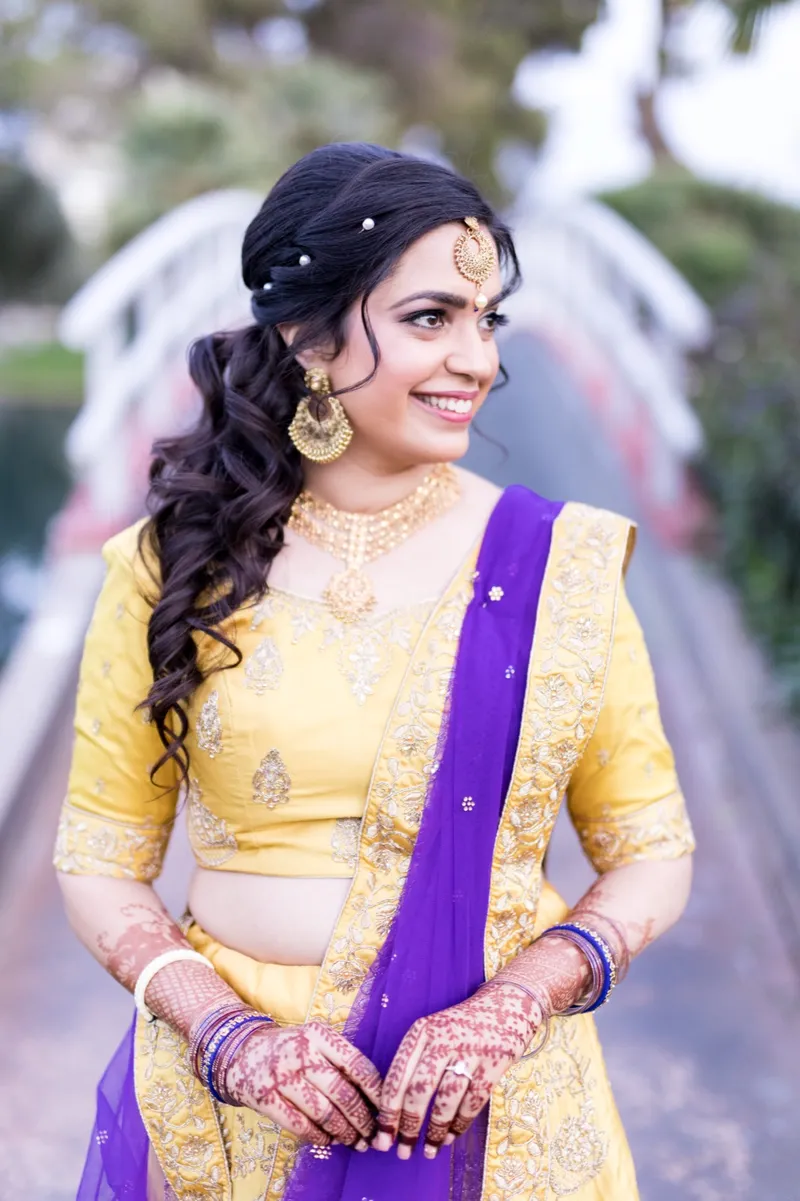 Bride in a yellow lehenga with gold jewelry and a maang tikka, standing on the path with the white bridge behind her