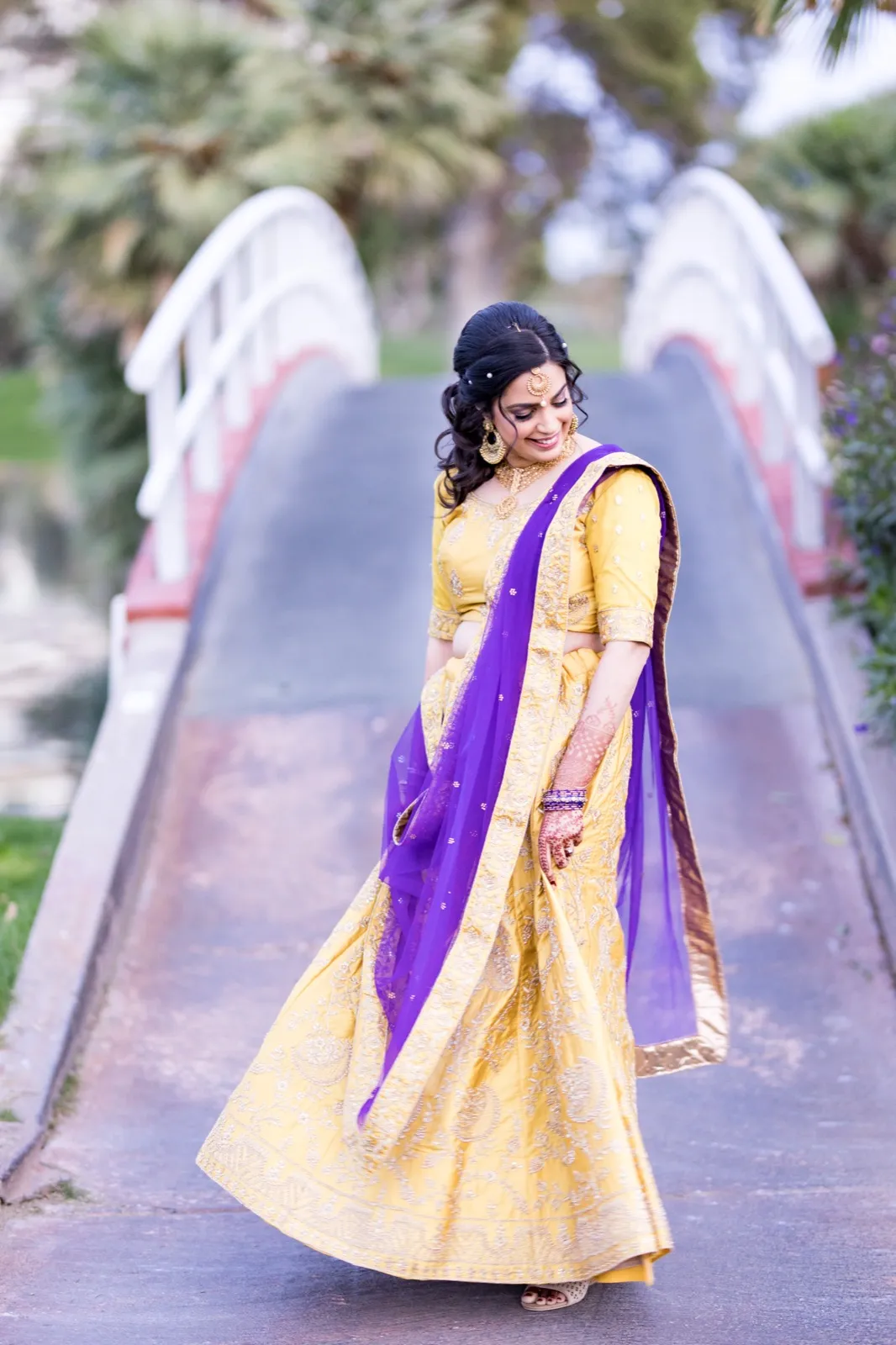 Bride in a yellow lehenga and purple dupatta walking down the white arched bridge over the lake