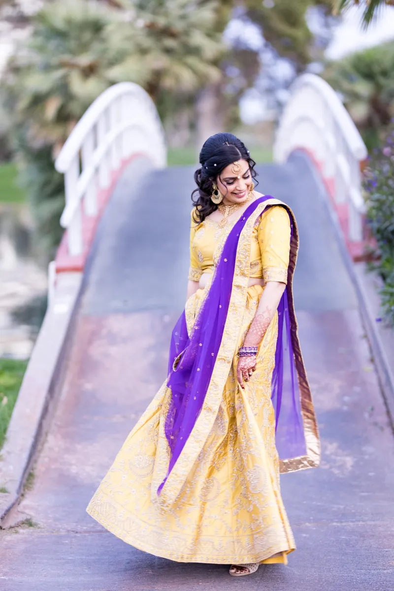 Bride in a gold embroidered yellow lehenga and purple dupatta walking down the white arched bridge over the water, palms behind her