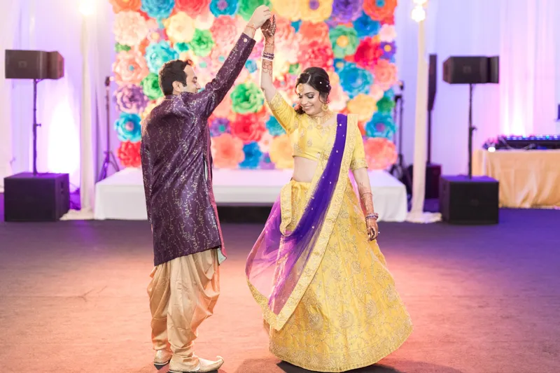 Groom in purple twirling the bride in yellow on the dance floor in front of a wall of bright paper flowers, with DJ speakers and a white stage behind