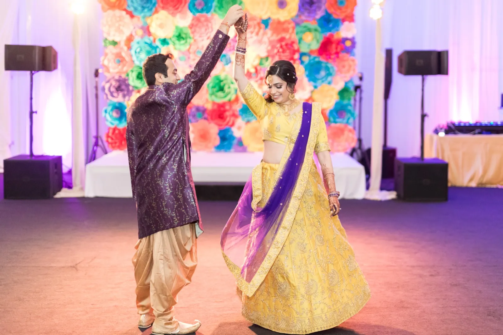 Groom twirls the bride on the dance floor in front of a wall of bright paper flowers