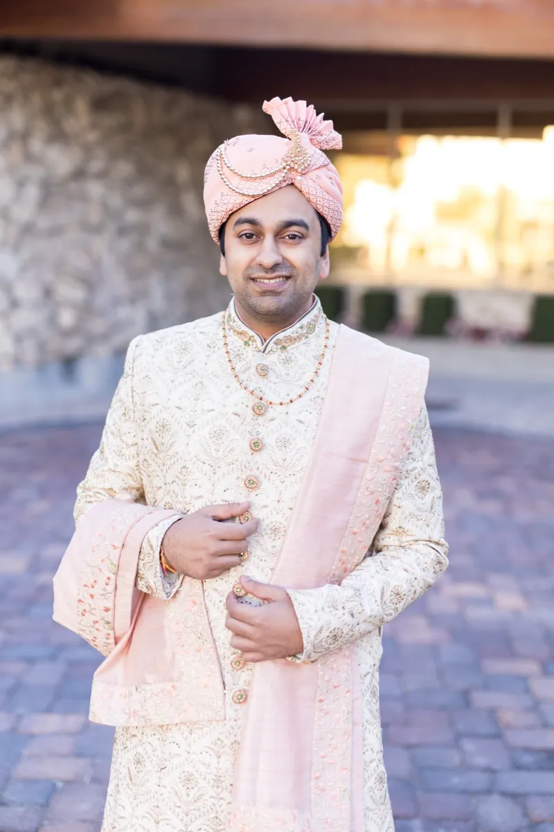 Groom standing on the entrance courtyard path in his sherwani and turban, the clubhouse rock wall behind him
