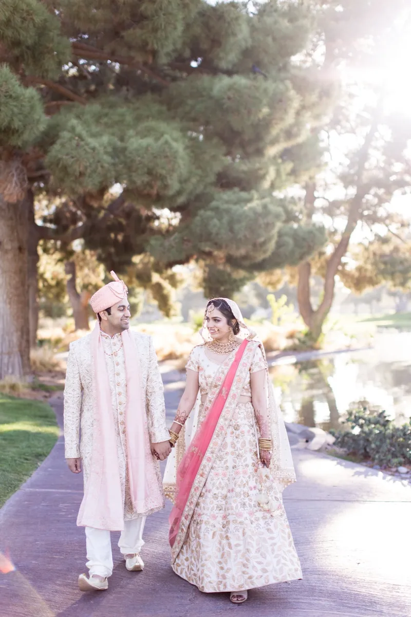 Couple in cream and coral walking hand in hand up the path under tall pines with low sun flaring through the branches