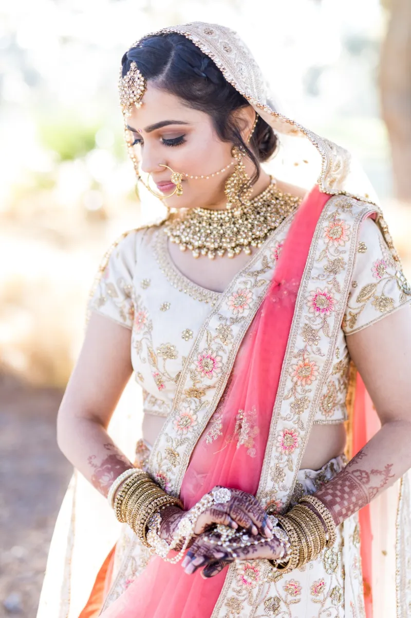 Bride in a cream and coral lehenga with layered gold jewelry, looking down and away from the camera