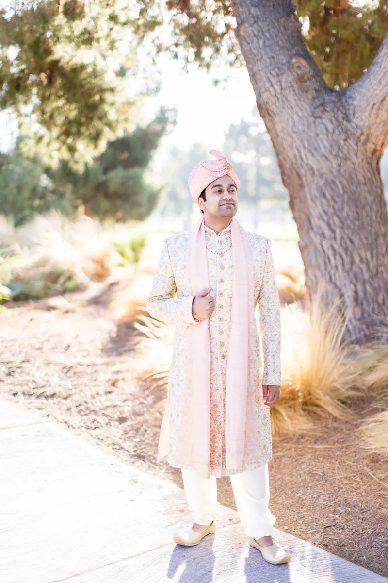 Groom in a cream sherwani standing under the bare branches of a tree at the edge of the course