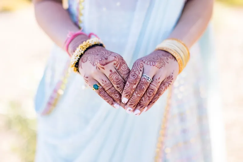 Close view of two henna covered hands and stacked pink and gold bangles resting against a pale blue outfit