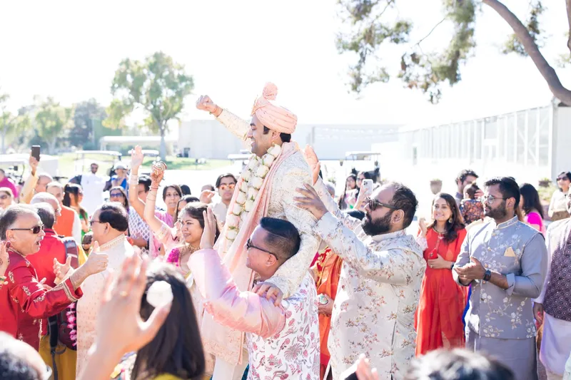 Groom in a peach turban and a white flower garland carried on shoulders with his fist raised, guests dancing around him and golf carts parked behind