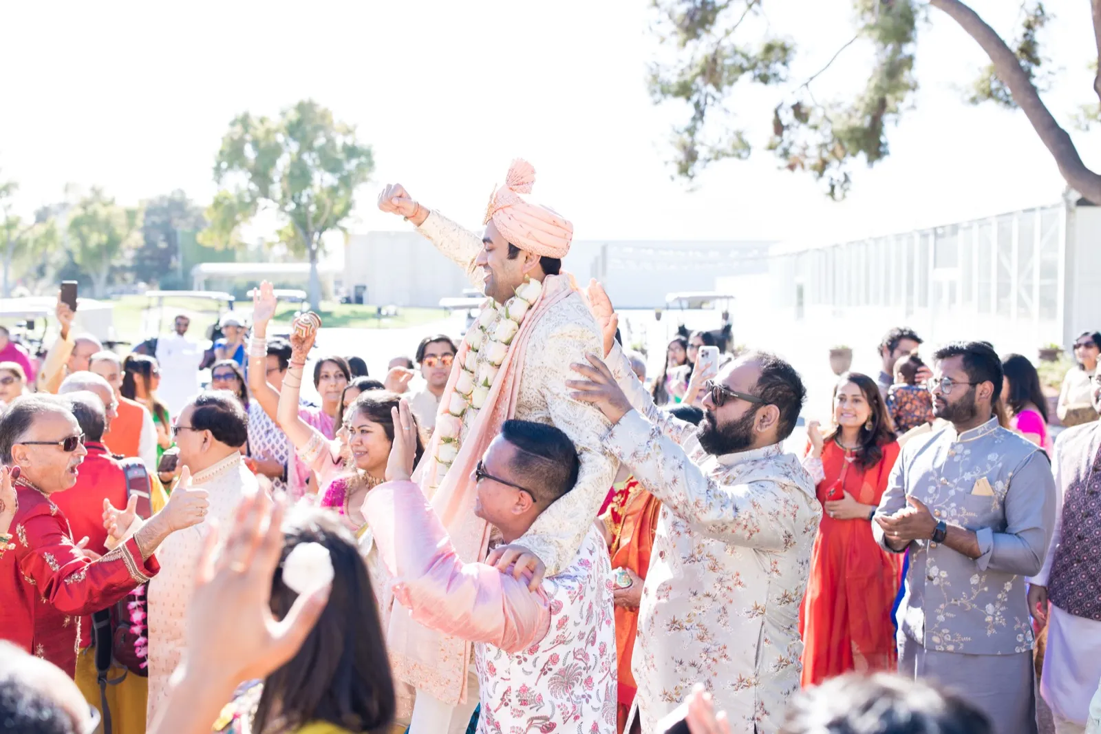 Groom in a peach turban and flower garland lifted on shoulders as guests dance around him