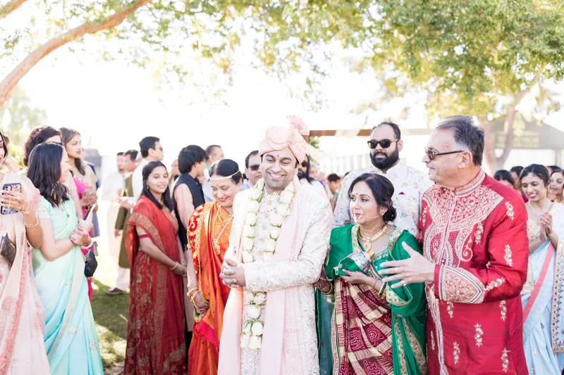 Couple in garlands surrounded by family under the trees, phones raised all around them
