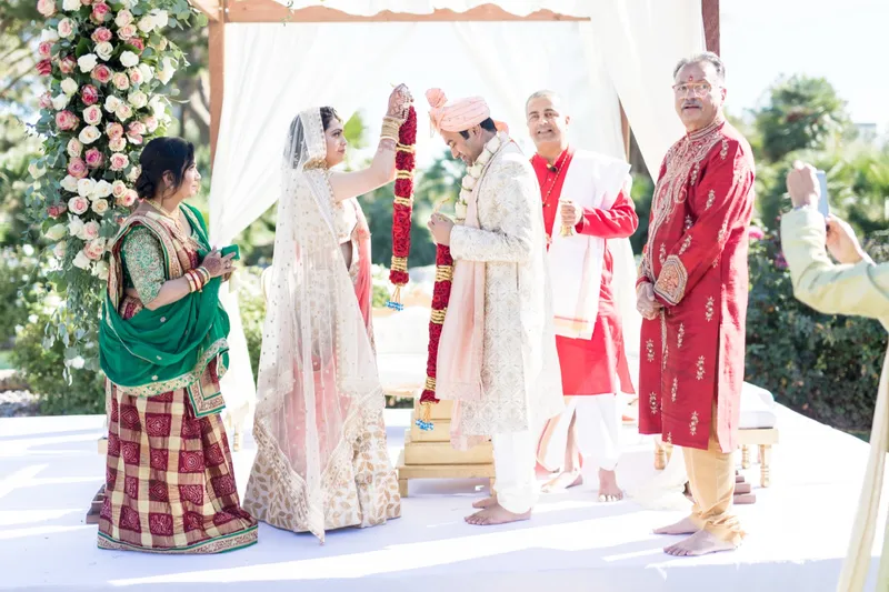 Bride lifting a red and gold garland over the groom's head at a rose covered mandap while family and the priest stand close by