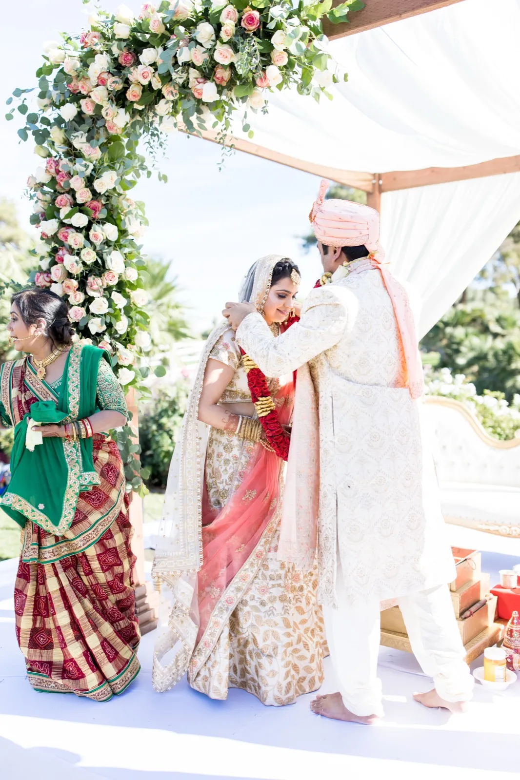 Bride and groom exchange red rose garlands under a flower covered mandap as a relative looks on