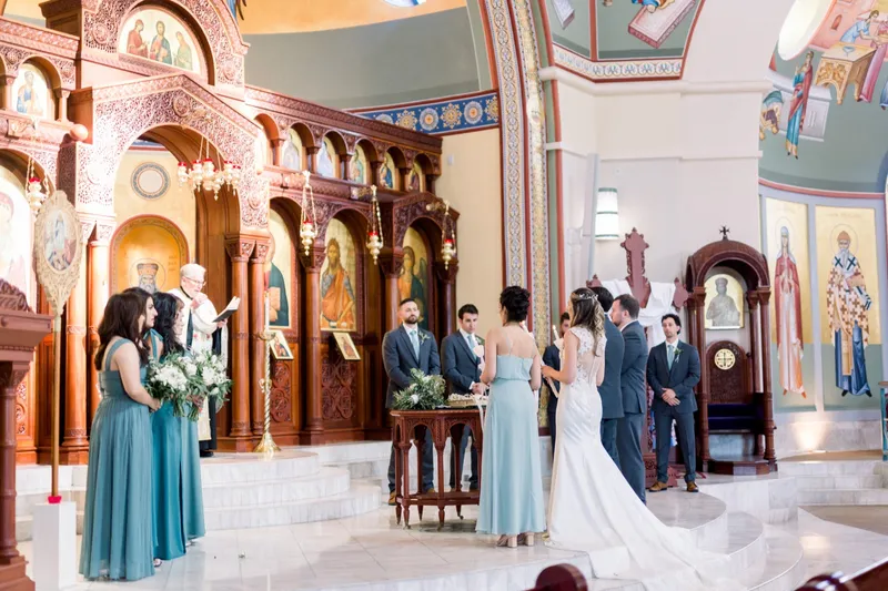 Bride and groom facing each other at the front of the church with the priest between them, bridesmaids in teal gowns standing along the marble step