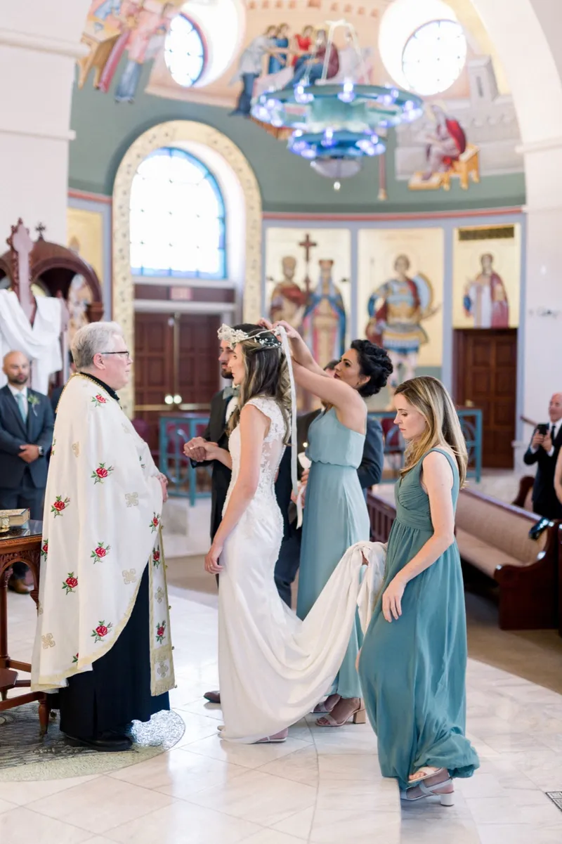 The priest in embroidered vestments beside the couple while a woman in a teal gown reaches up to move the linked wedding crowns above their heads