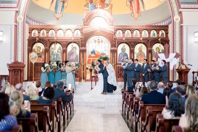 Wide view of the church from the back of the aisle, the couple kissing at the top of the marble steps in front of the carved wooden iconostasis while their wedding party stands on both sides and guests fill the pews