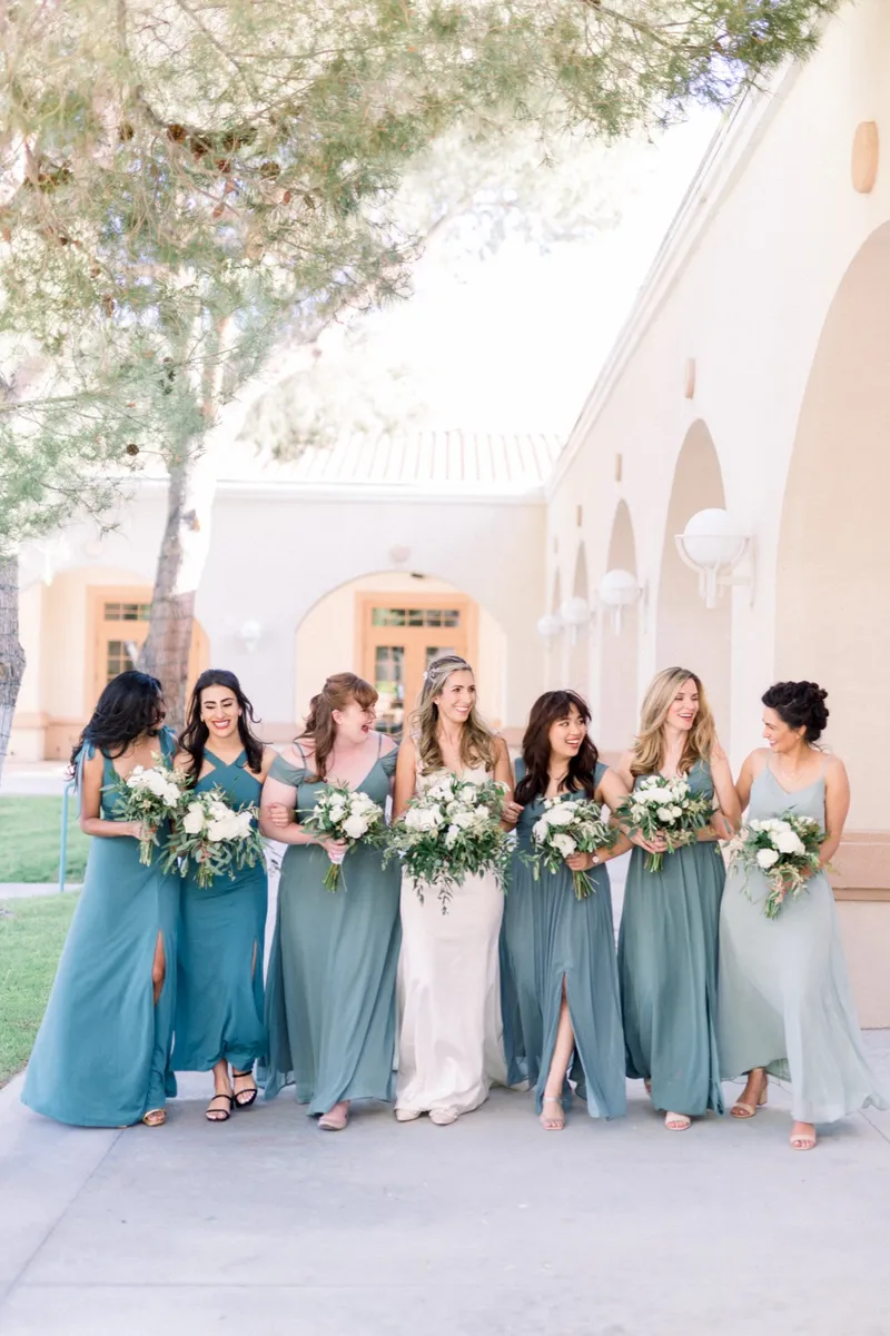Bride and her bridesmaids laughing together under a tree, the arched walkway and red tile roof of the church behind them