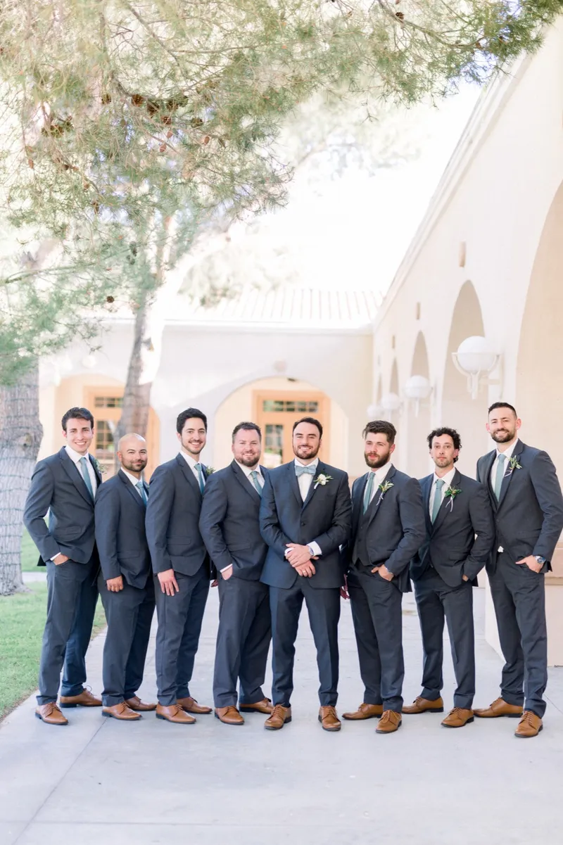 Groomsmen in slate suits and sage ties standing in a long line on the concrete in front of the church arches