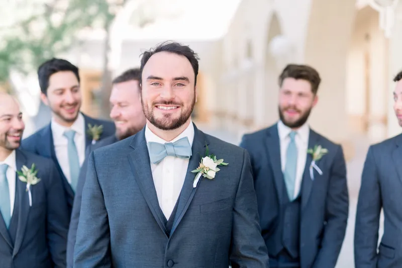 Groom grinning at the camera in a pale blue bow tie with his groomsmen fanned out behind him
