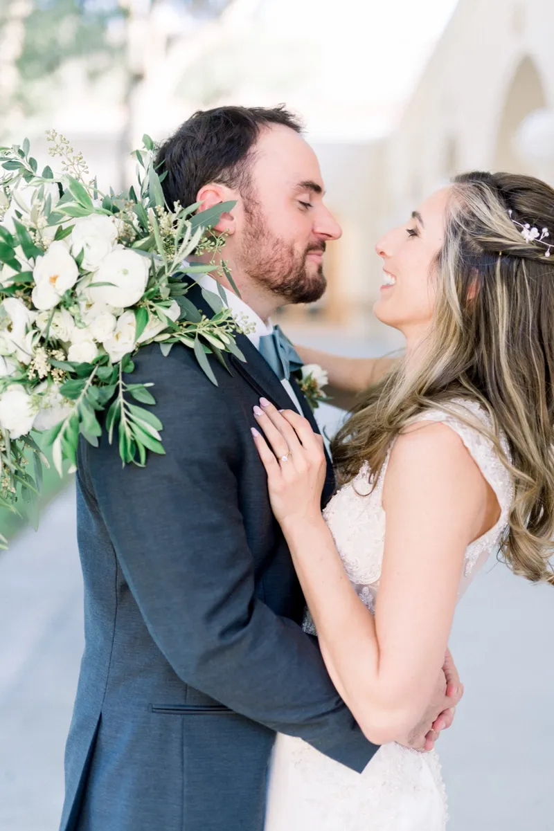 Bride and groom leaning close together with her hand on his chest and her white and olive bouquet up at his shoulder