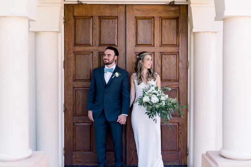 Bride and groom standing in the tall carved wooden doorway of the church, pale round columns on both sides of them