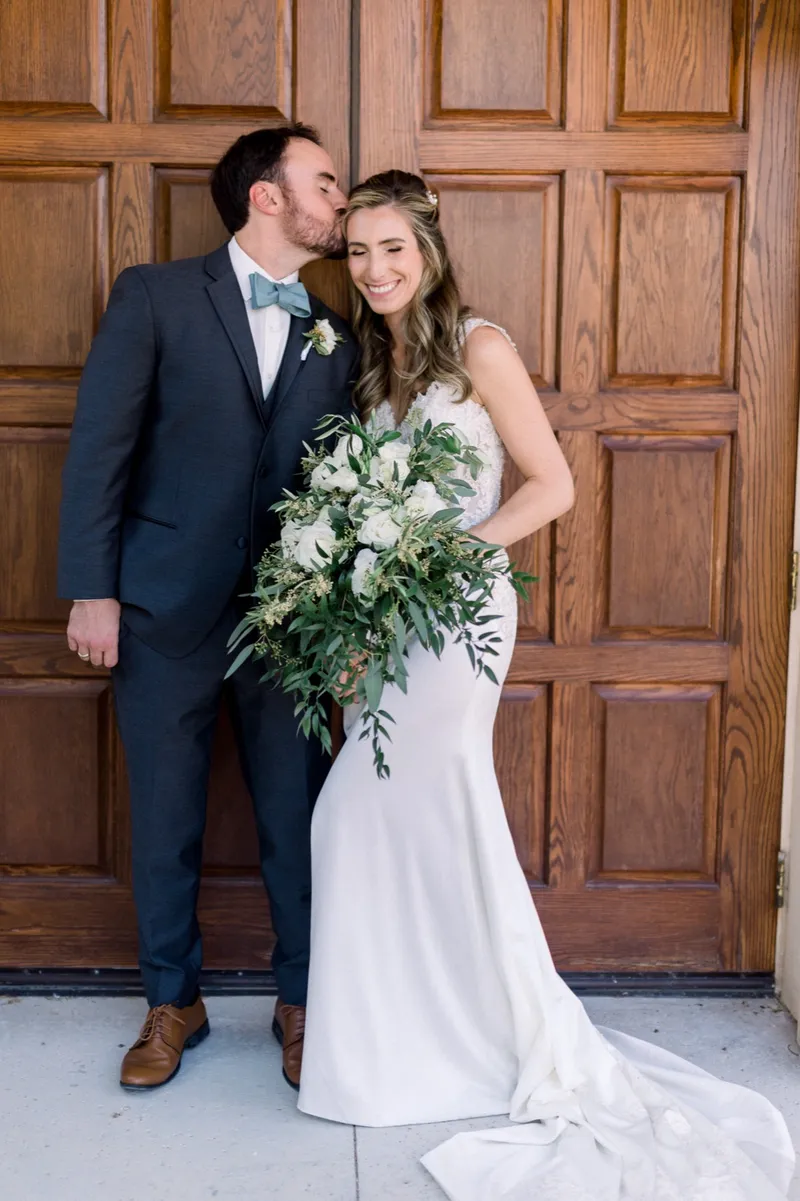 Groom kissing the top of the bride's head in front of the heavy carved doors, her train pooled on the ground beside them