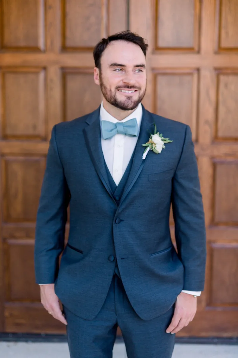 Groom standing on his own in a slate suit and pale blue bow tie, an olive and white boutonniere on his lapel