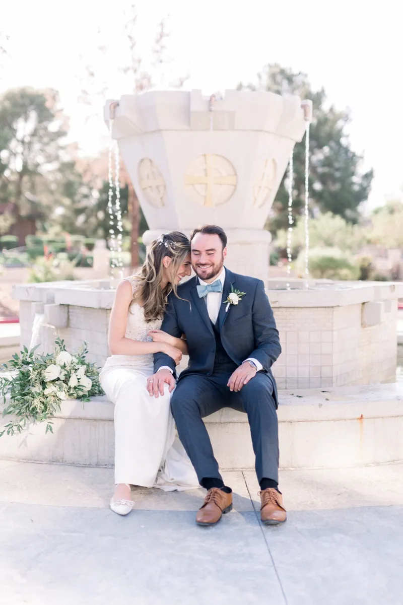 Bride and groom sitting together on the rim of the stone fountain outside the church, an Orthodox cross carved into the fountain wall behind them