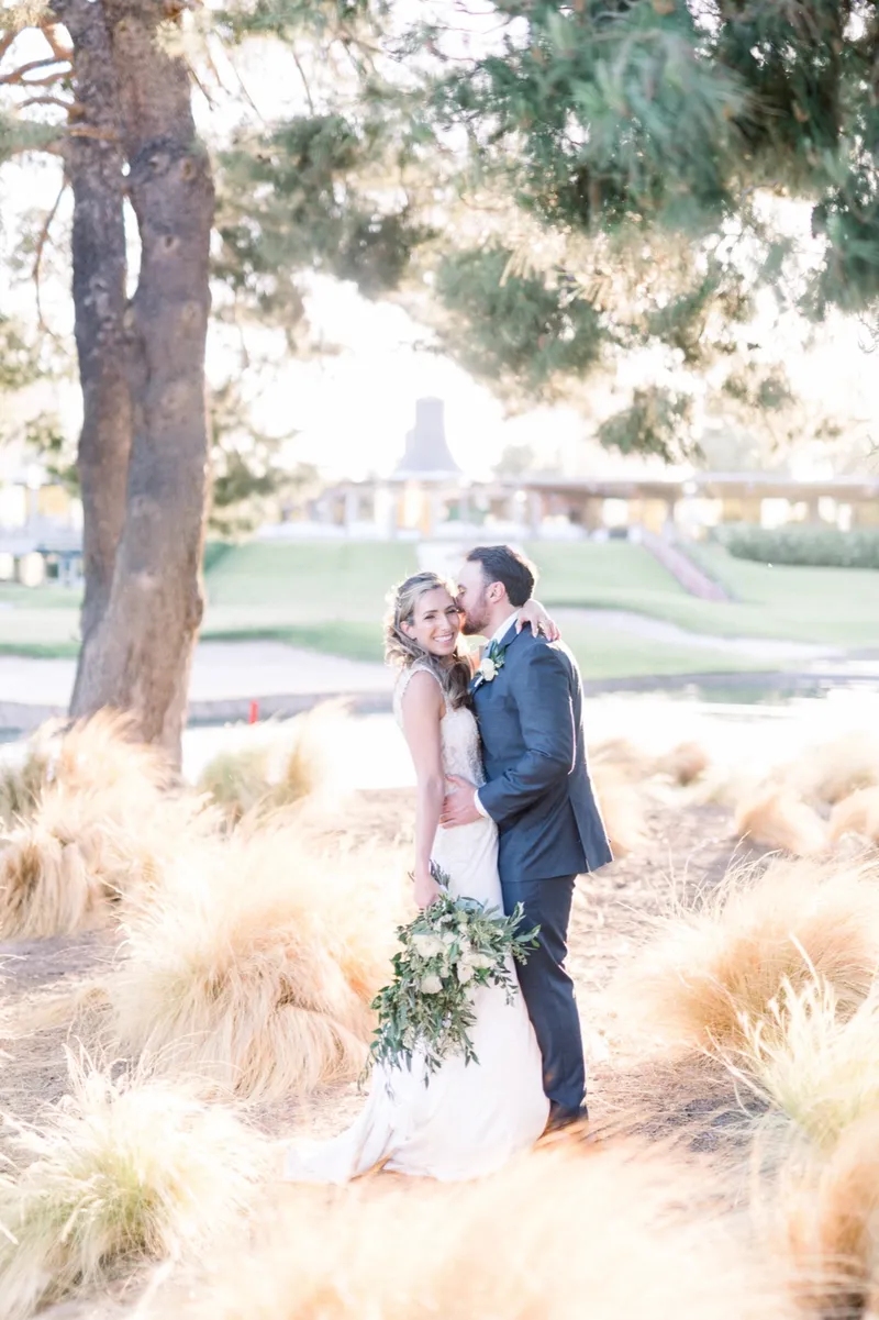 Groom in a navy suit kissing his bride's cheek as they stand in tall golden ornamental grasses with the clubhouse behind them
