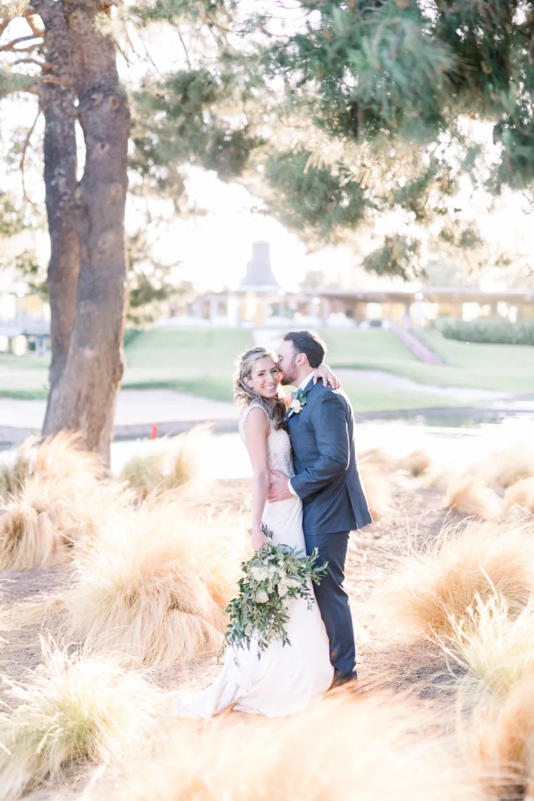 A groom kisses his bride’s cheek among tall golden grasses on a golf course in warm late light.