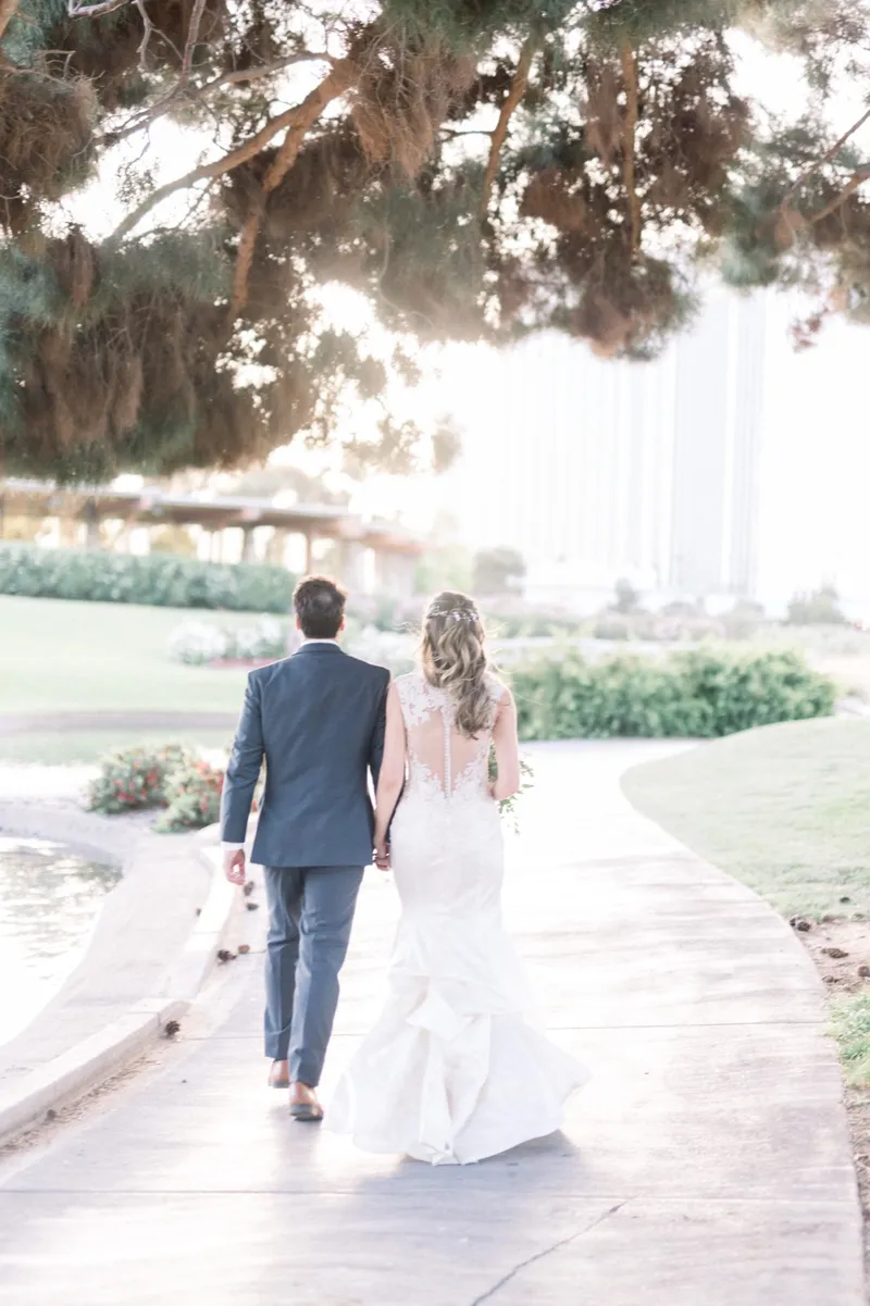 Couple walking away hand in hand down a path beside the water, the bride's train trailing behind her under mature trees