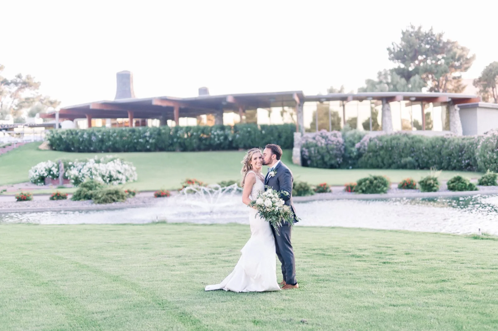 Bride and groom kissing in the middle of a striped mown lawn, with clipped hedges, a fountain in the water and the long low clubhouse behind them
