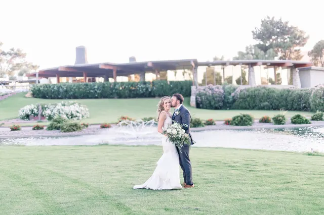 Bride and groom kissing in the middle of a striped mown lawn, with clipped hedges, a fountain in the water and the long low clubhouse behind them