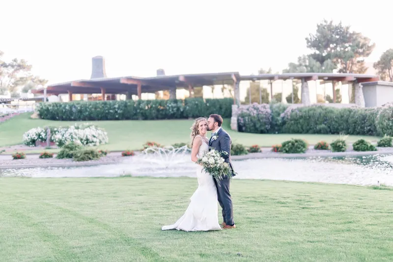 Groom kisses his bride's cheek on the lawn, the lake fountain and low clubhouse behind them