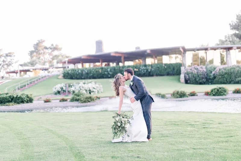 Groom in a grey suit dipping his bride into a kiss on the lawn, her bouquet swinging low, the fountain and clubhouse behind them