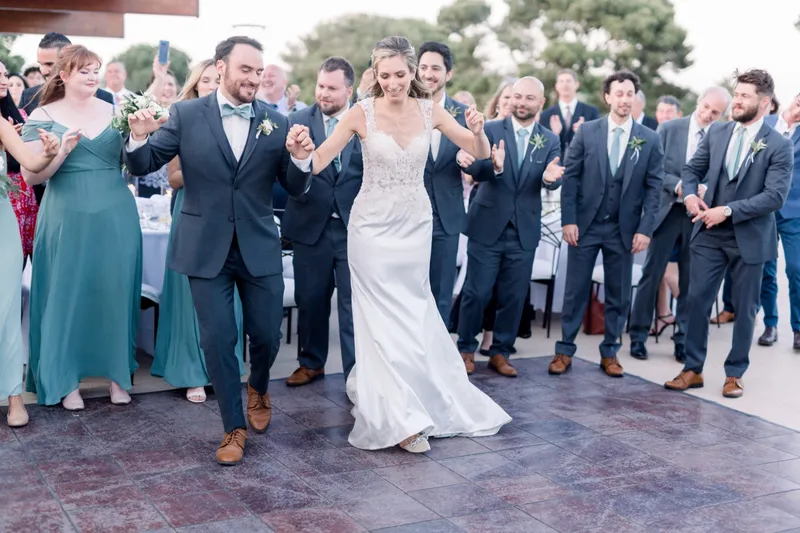 Couple dancing onto the tiled terrace floor for their entrance while the wedding party in slate suits and sage dresses cheers