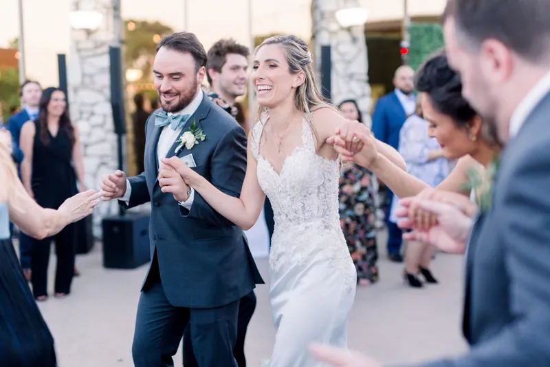 Couple dancing hand in hand in a ring of guests at dusk, the terrace stone columns and lamps lit behind them