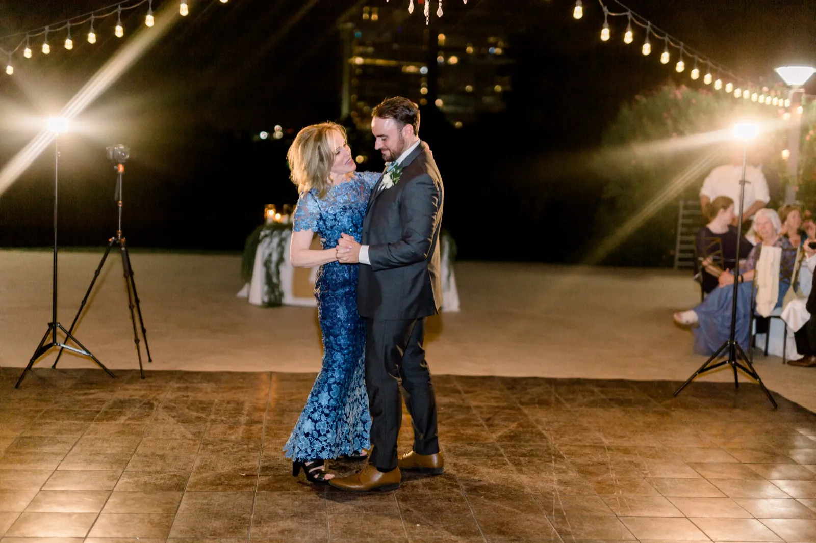 A groom dances with an older woman in a blue lace gown on an outdoor floor under string lights at night