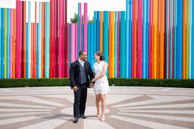 Couple walking hand in hand across a paved circle in front of a long wall of tall colored pipes, him in a black suit and white bow tie, her in a short white lace dress and pink heels