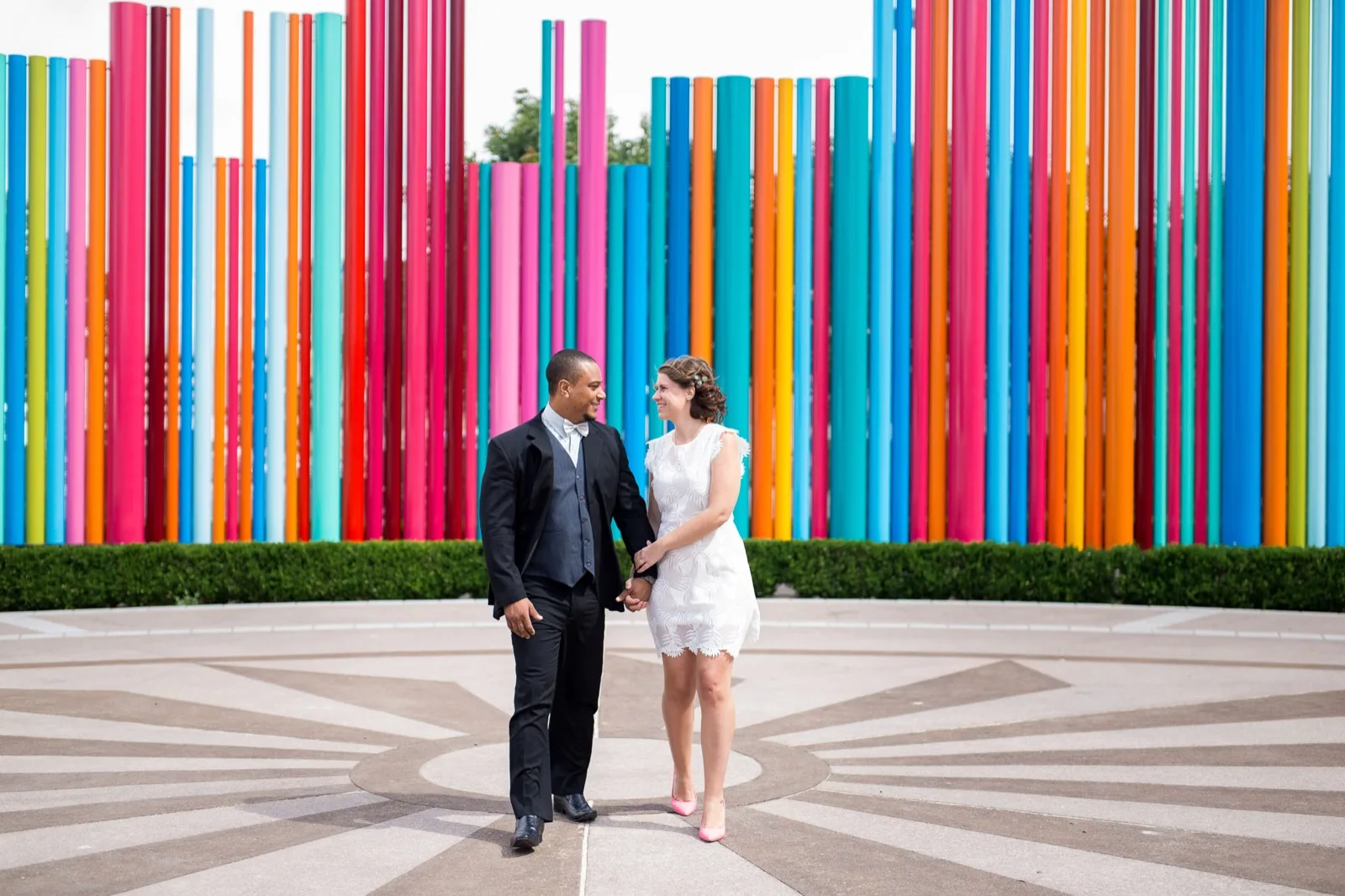 Couple walking hand in hand across a paved circle in front of a long wall of tall colored pipes, him in a black suit and white bow tie, her in a short white lace dress and pink heels