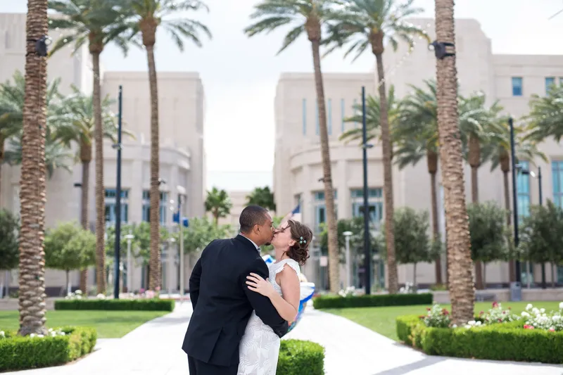 Couple kissing on a strip of green lawn between palm trees, low hedges and the stone buildings framing them