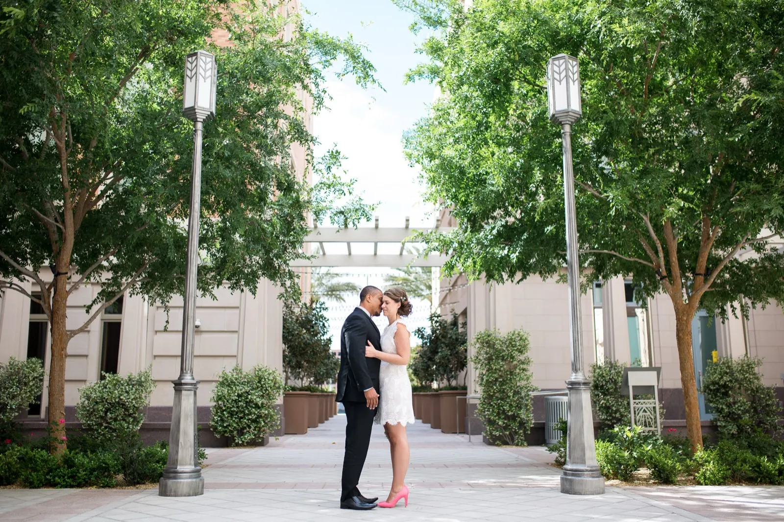Couple standing forehead to forehead on flat paving between two tall art deco lampposts and leafy trees
