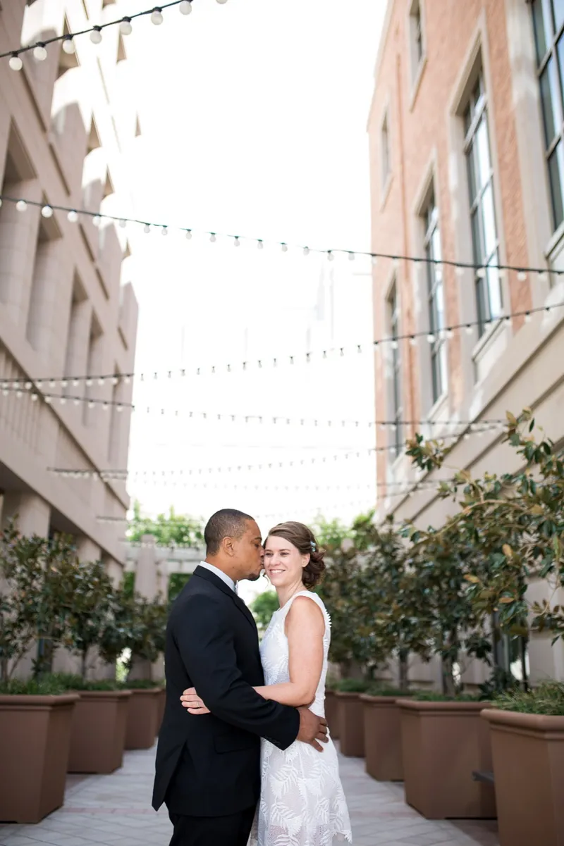 Couple standing close on the walkway under the strung lights, her short white lace dress against his black suit