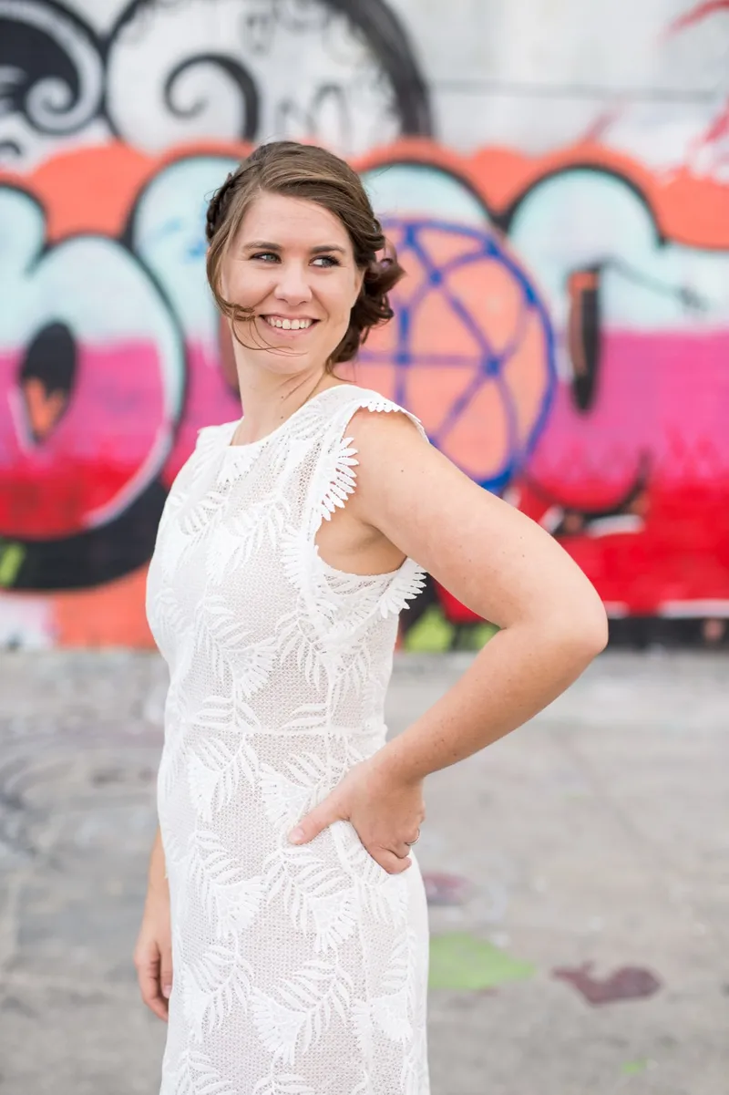 Bride in a short white lace dress standing with one hand on her hip against the same bright graffiti wall