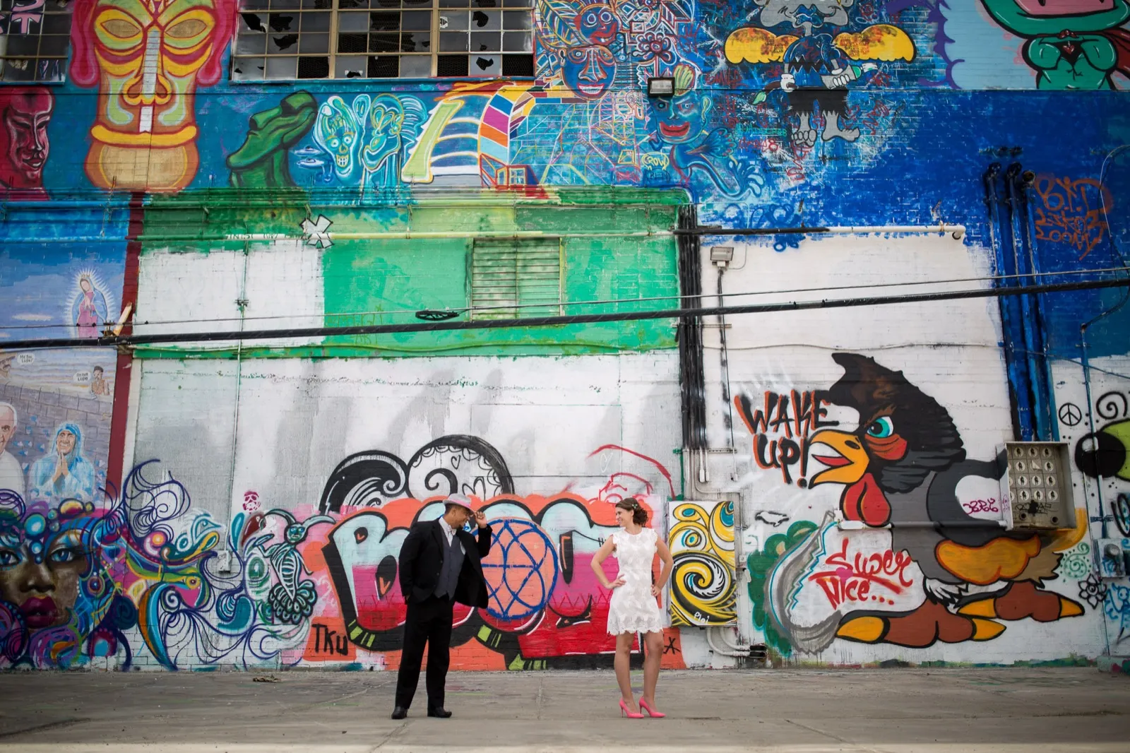 Groom tipping his straw fedora to his bride from the far end of a block long wall painted with tiki faces and a rooster