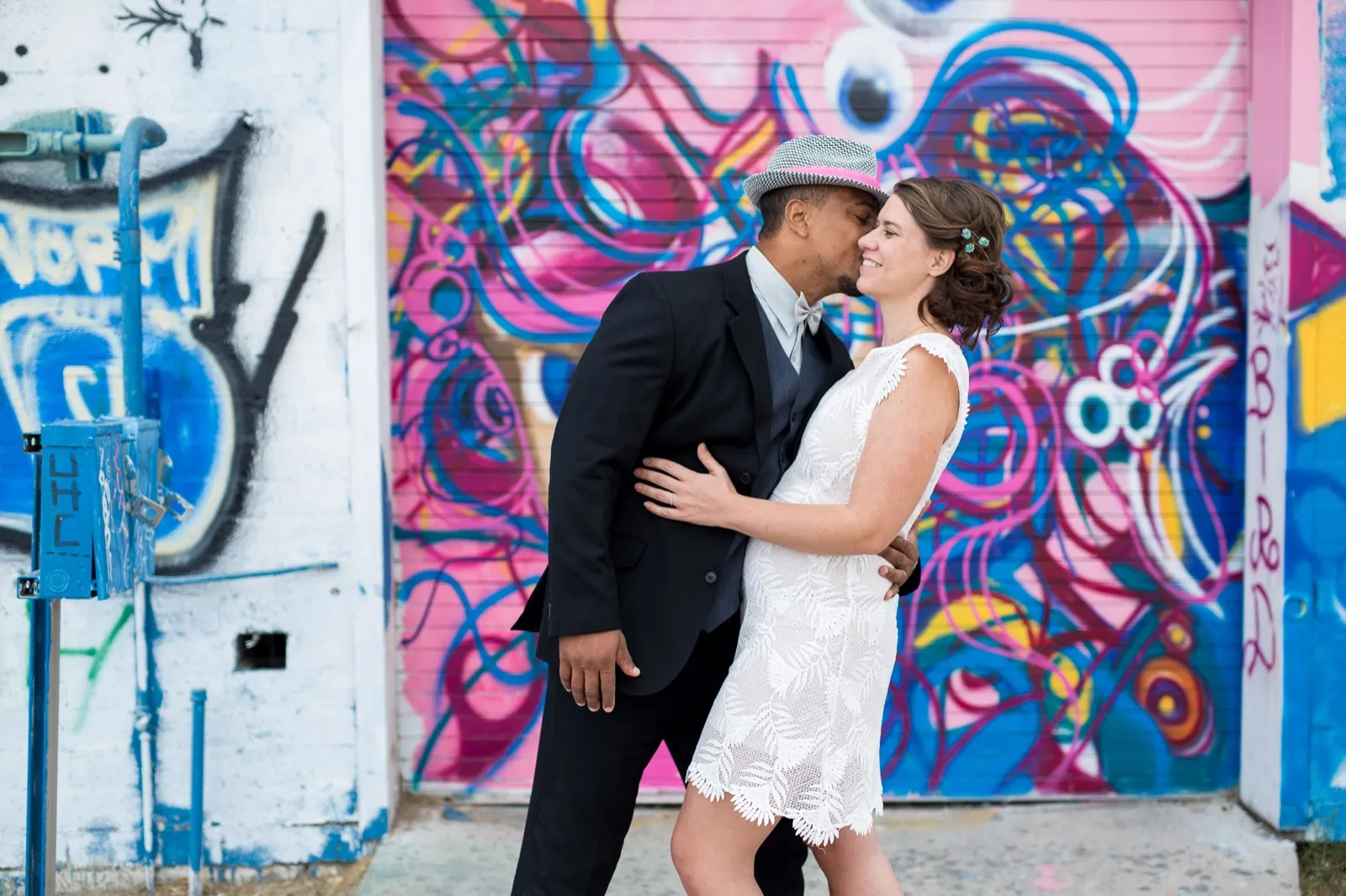 Groom in a black suit and straw fedora kissing his bride’s cheek in front of a pink and blue mural in downtown Las Vegas