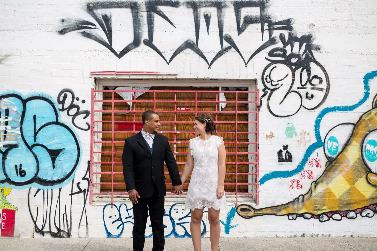 Couple holding hands in front of a white brick warehouse front covered in graffiti, a red barred window behind them