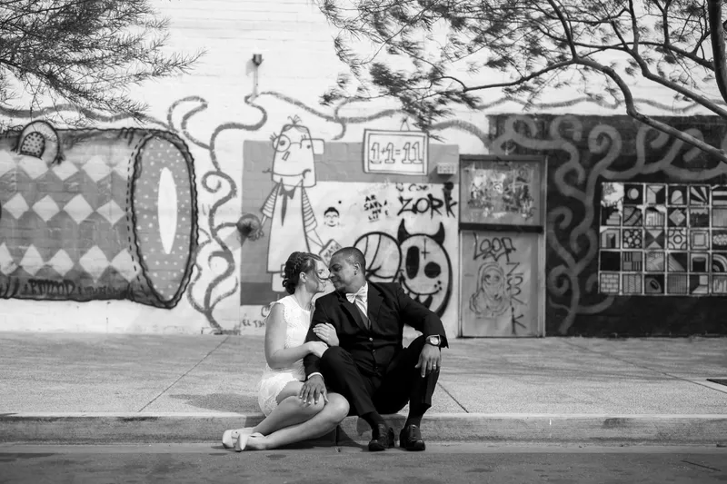 Black and white photo of the couple sitting together on the curb with a wall of cartoon doodles behind them