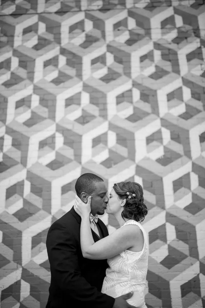 Black and white photo of the couple kissing in front of a wall painted in a repeating cube pattern